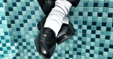 Pernas cruzadas com sapatos pretos e meias brancas diante de um fundo de azulejos azuis, em composição de estilo retrô e urbano. Os sapatos lembram o estilo do Michael Jackson.
