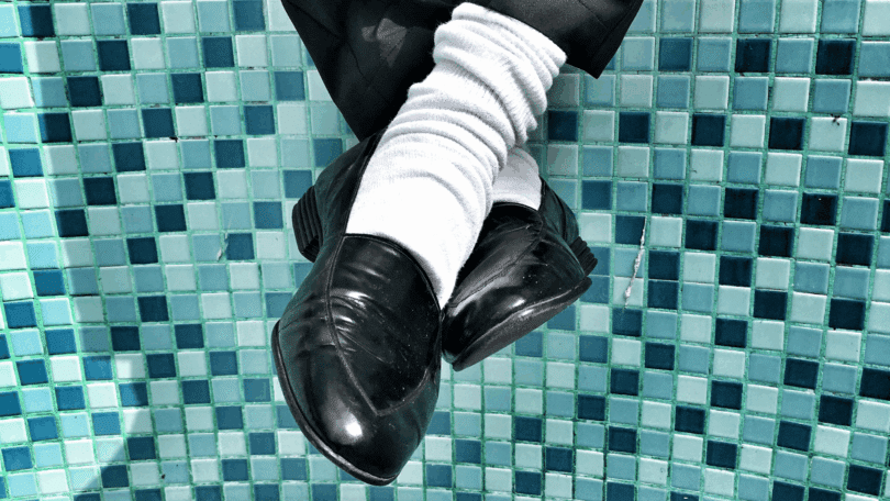 Pernas cruzadas com sapatos pretos e meias brancas diante de um fundo de azulejos azuis, em composição de estilo retrô e urbano. Os sapatos lembram o estilo do Michael Jackson.
