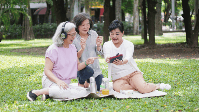 Três mulheres idosas sentadas sobre uma toalha em um parque arborizado observam um celular e sorriem; uma usa fones de ouvido, enquanto garrafas e lanches estão ao lado delas na grama.