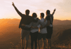 Grupo de amigos abraçados contempla o pôr do sol em uma paisagem montanhosa, celebrando um momento de união e liberdade.