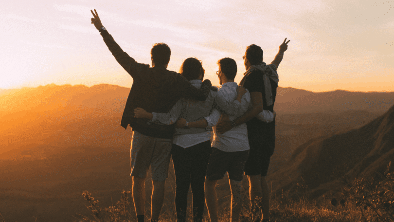 Grupo de amigos abraçados contempla o pôr do sol em uma paisagem montanhosa, celebrando um momento de união e liberdade.