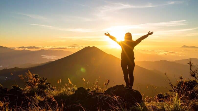 Imagem de uma mulher em cima de um morro, olhando para uma linda montanha. Ela está de braços abertos, recebendo a luz do Sol.
