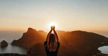Imagem de uma mulher com os braços erguidos olhando para a luz do sol em frente a uma montanha.