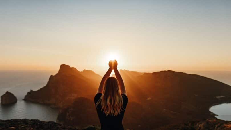 Imagem de uma mulher com os braços erguidos olhando para a luz do sol em frente a uma montanha.