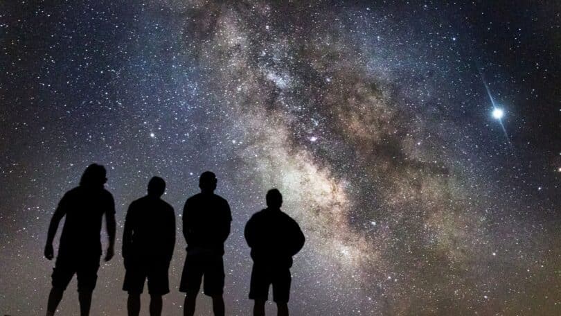 Imagem de um céu cósmico repleto de estrelas, com a silhueta de 4 pessoas ao centro olhando para o infinito, simbolizando a origem da existência, consciência e a eterna busca por respostas sobre o que existia antes do nascimento.