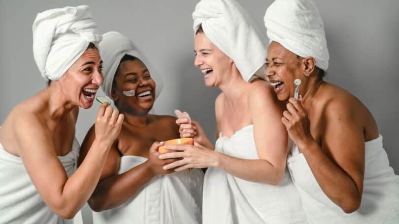 Imagem de quatro mulheres 50+ participando de um spa. Elas estão felizes, sorridentes enroladas em toalhas de banho na cor branca.