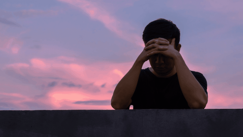Homem com as mãos na cabeça reza diante de um céu ao entardecer.