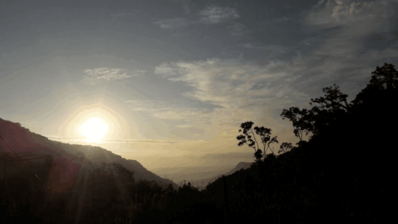 Paisagem ao amanhecer com sol no horizonte, montanhas ao fundo e árvores em silhueta.