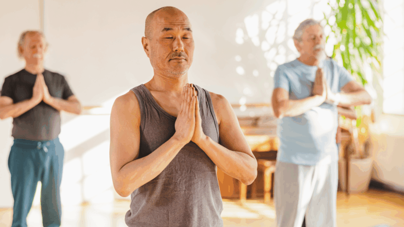 Grupo de pessoas pratica meditação em ambiente iluminado, com mãos unidas e olhos fechados.