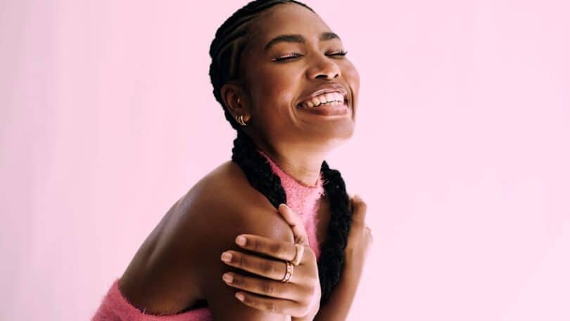 Imagem de fundo rosa e em destaque uma mulher negra se abraçando e sorrindo, simbolizando o conceito de auto amor.