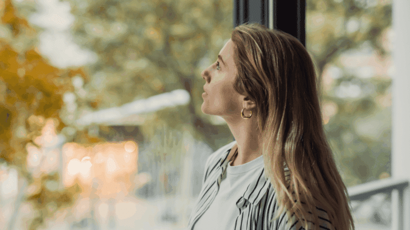 Mulher olhando pela janela com expressão contemplativa, tendo árvores desfocadas ao fundo e iluminação natural suave.
