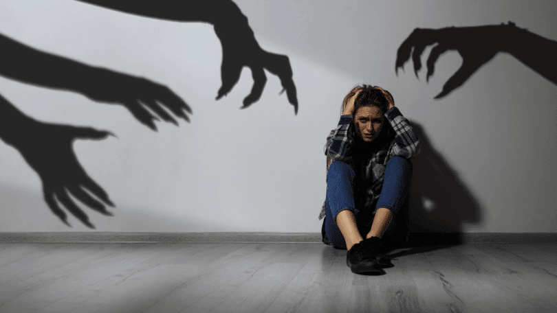 Mulher encolhida e angustiada no chão enquanto sombras de mãos ameaçadoras se projetam na parede ao seu redor.