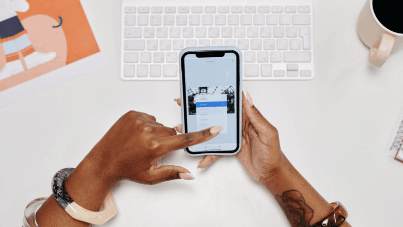 Mulher ajustando configurações de edição de imagem no celular sobre uma mesa com teclado, café e materiais de trabalho.