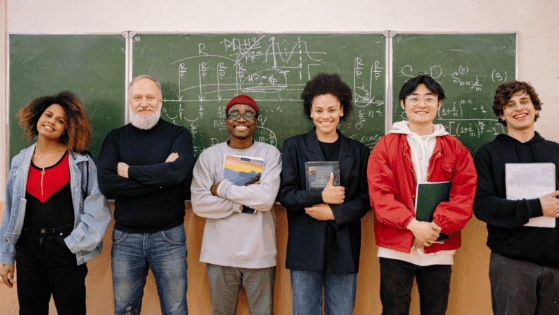 Grupo de professor e estudantes sorrindo em frente a um quadro com fórmulas matemáticas, segurando cadernos e livros.
