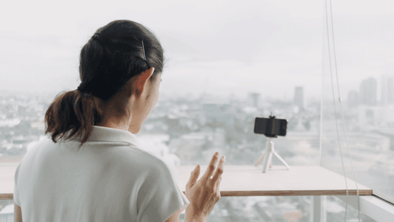 Mulher gravando vídeo com celular em tripé diante de uma janela com vista para a cidade.