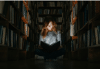 Imagem de uma pessoa sentada diante de uma grande estante de livros, em um ambiente pouco iluminado. A composição transmite a ideia de busca pelo conhecimento, questionamento e expansão da consciência.