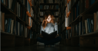 Imagem de uma pessoa sentada diante de uma grande estante de livros, em um ambiente pouco iluminado. A composição transmite a ideia de busca pelo conhecimento, questionamento e expansão da consciência.