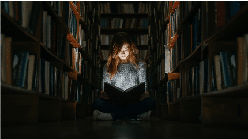 Imagem de uma pessoa sentada diante de uma grande estante de livros, em um ambiente pouco iluminado. A composição transmite a ideia de busca pelo conhecimento, questionamento e expansão da consciência.