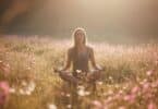 Imagem de uma mulher meditando, sentanda emum campo florido com natureza e uma luz espiritual ao fundo.