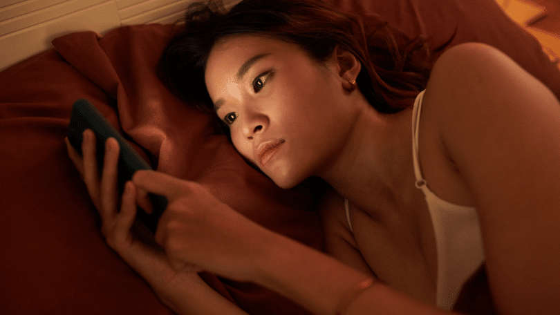 Imagem de uma mulher sozinha, deitada sobre a sua cama, olhando para o celular. A foto simboliza o paradoxo de estar conectado e, ainda assim, sentir-se sozinho.