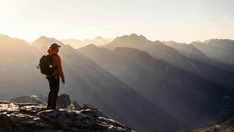 Homem com mochila observa uma paisagem montanhosa do alto de um pico.