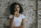 Mulher pensativa, com a mão no queixo, diante de uma parede de tijolos.