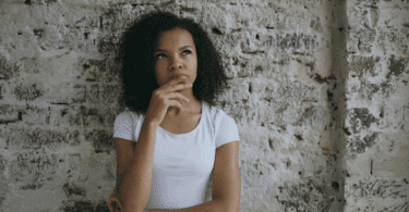 Mulher pensativa, com a mão no queixo, diante de uma parede de tijolos.