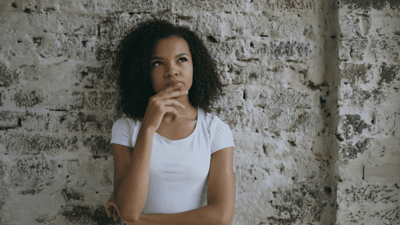 Mulher pensativa, com a mão no queixo, diante de uma parede de tijolos.