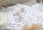 Imagem de um mapa astral sobre uma mesa, uma taça com uma vela branca acesa e 3 pedras brancas com símbolos do zodíaco.