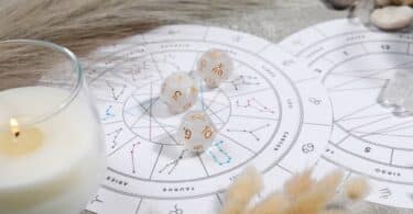 Imagem de um mapa astral sobre uma mesa, uma taça com uma vela branca acesa e 3 pedras brancas com símbolos do zodíaco.