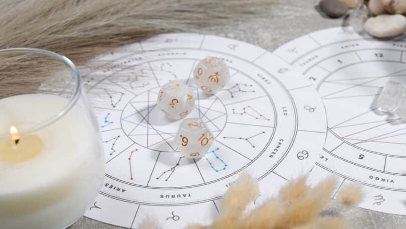 Imagem de um mapa astral sobre uma mesa, uma taça com uma vela branca acesa e 3 pedras brancas com símbolos do zodíaco.