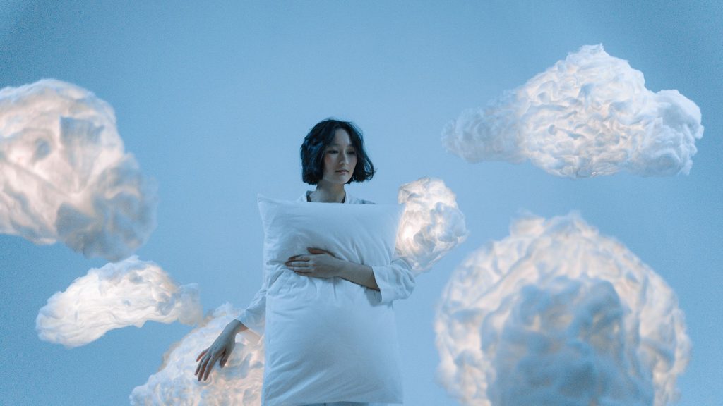 Imagem de uma mulher segurando um travesseiro branco e ao lado dela, várias nuvens simbolizando os seus sonhos.