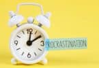 Imagem de fundo amarelo e em destaque um despertador branco e a palavra PROCRASTINAÇÃO escrita em inglês - PROCRASTINATION.