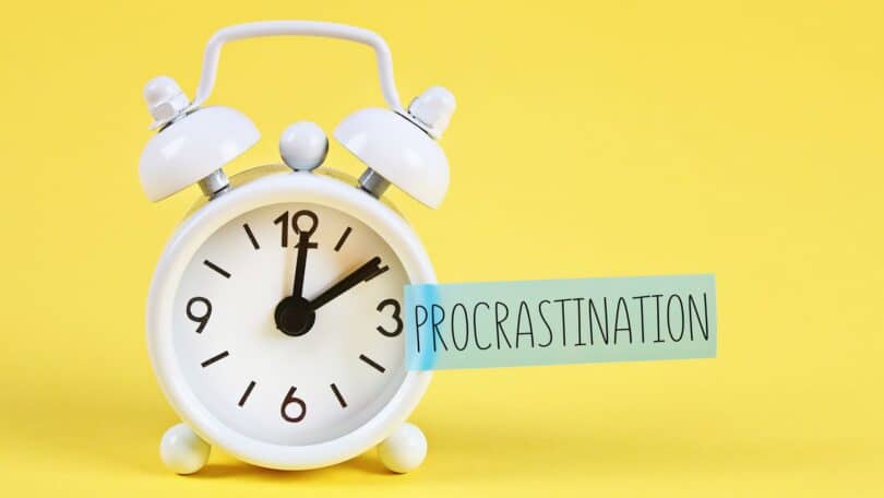 Imagem de fundo amarelo e em destaque um despertador branco e a palavra PROCRASTINAÇÃO escrita em inglês - PROCRASTINATION.