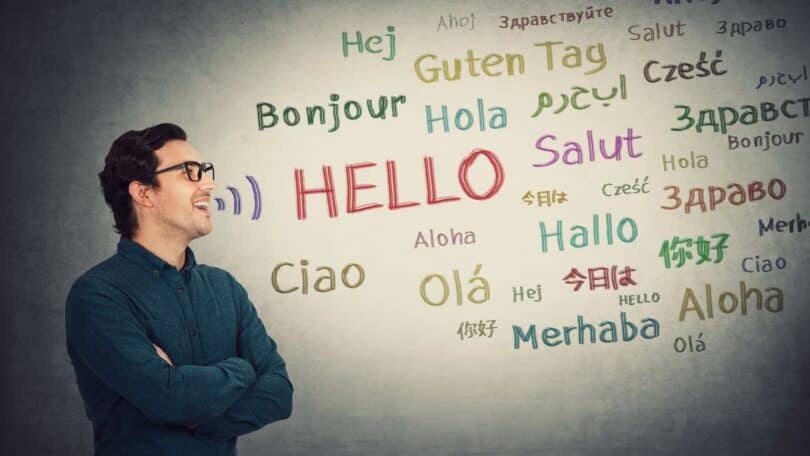 Imagem de um quadro branco e um homem ao lado. No quadro, existe a palavra Olá, escrita em vários idiomas, simbolizando o conceito de tradução e comunicação.