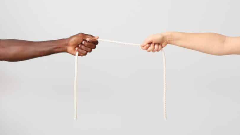 Imagem de dois braços, um de uma pessoa negra e outro de uma pessoa branca, segurando uma corda esticada entre eles, simbolizando a xenofobia, o preconceito e a divisão entre pessoas de diferentes origens.