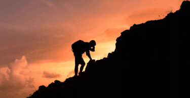 Homem escalando uma formação rochosa ao pôr do sol, em silhueta.