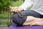 Mulher meditando ao ar livre, sentada sobre um tapete e segurando um colar de contas.
