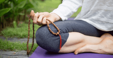 Mulher meditando ao ar livre, sentada sobre um tapete e segurando um colar de contas.