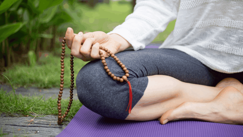 Mulher meditando ao ar livre, sentada sobre um tapete e segurando um colar de contas.