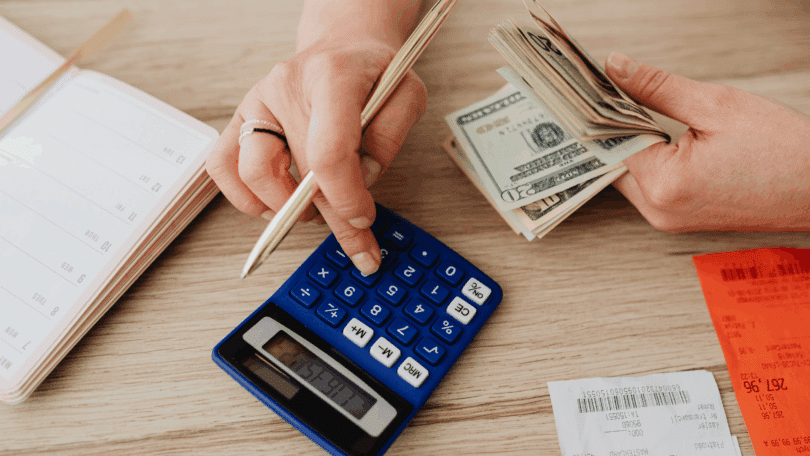 Pessoa calculando despesas e organizando as finanças com dinheiro em dólares, calculadora, agenda e recibos sobre uma mesa.