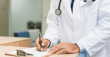 Médico de jaleco branco preenche um formulário em uma prancheta durante atendimento em consultório.