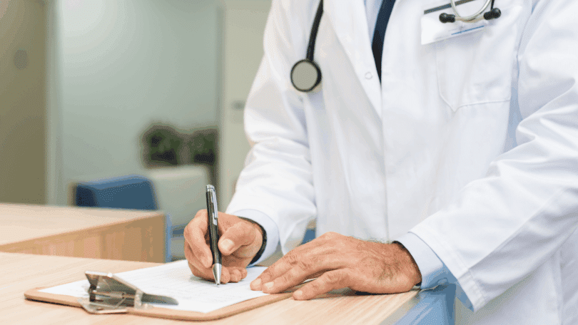 Médico de jaleco branco preenche um formulário em uma prancheta durante atendimento em consultório.