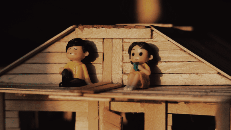 Duas bonecas sentadas em uma casinha de madeira iluminada por uma luz quente, representando uma cena familiar em miniatura.