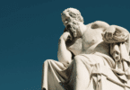 Escultura de mármore de Sócrates sentado, com a mão apoiada no rosto em uma pose reflexiva, sob um céu azul.