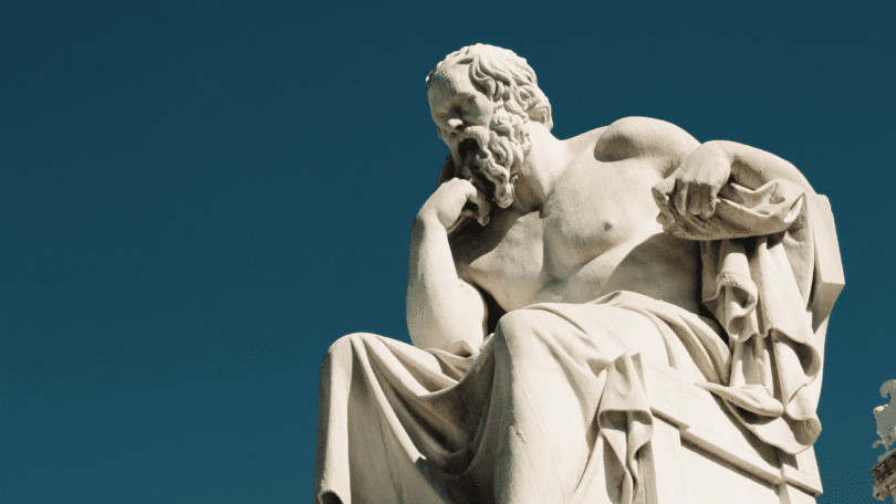Escultura de mármore de Sócrates sentado, com a mão apoiada no rosto em uma pose reflexiva, sob um céu azul.